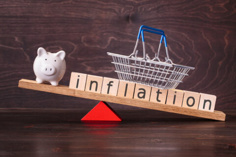 Inflation spelled in letters with shopping basket and piggy bank on seesaw against dark background